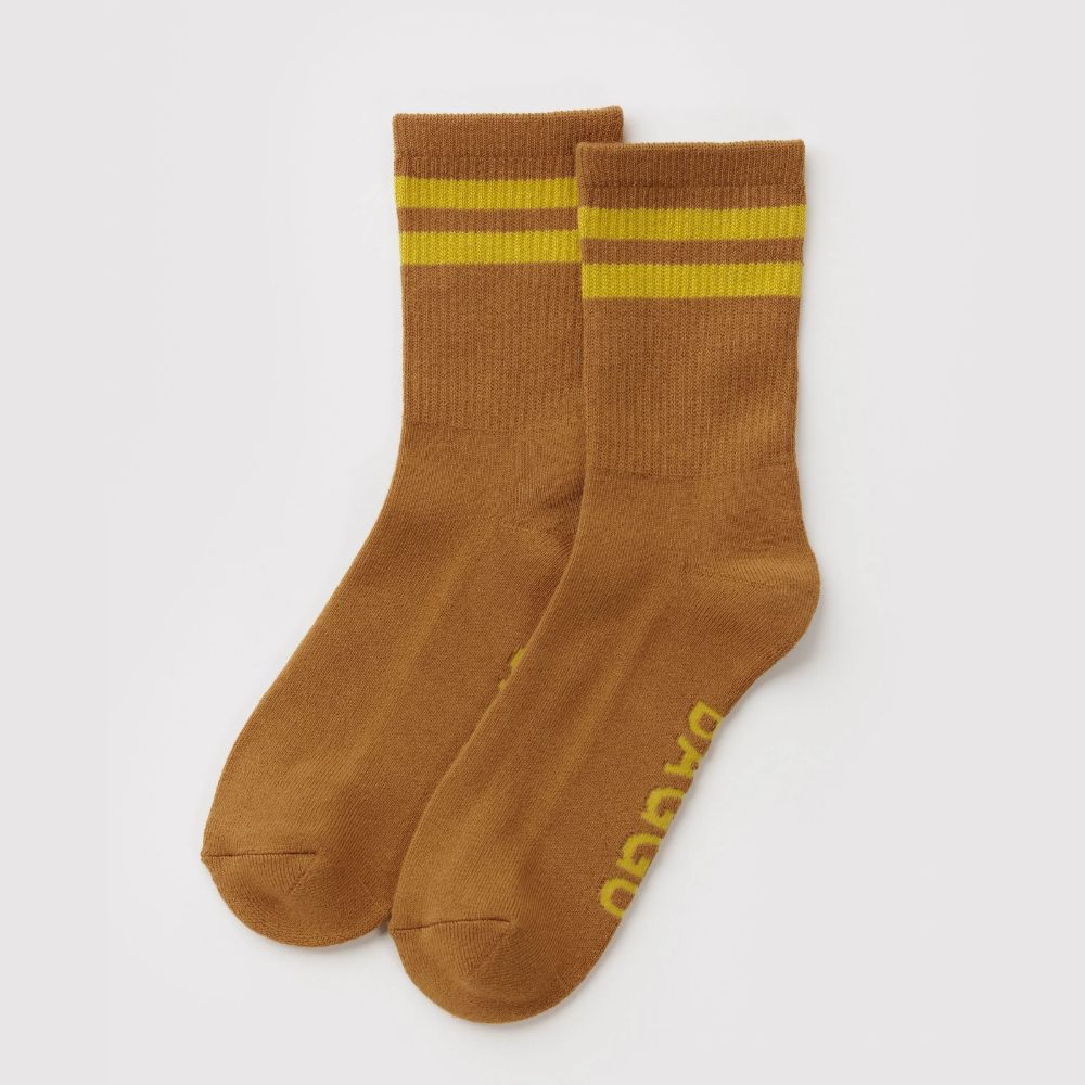 A pair of brown crew socks with two yellow stripes near the cuff and yellow branding printed on the sole, displayed on a light gray background.