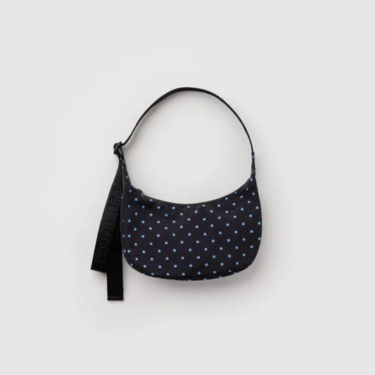 A crescent-shaped shoulder bag with a navy blue base and light blue polka dots, featuring a black adjustable strap, shown against a plain light gray background.