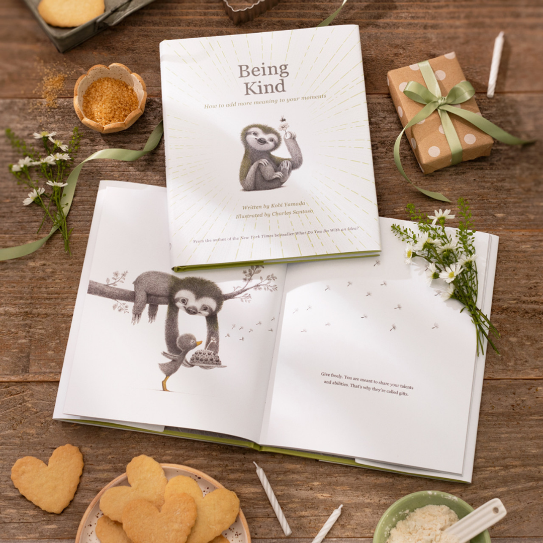 2 copies of "Being Kind" sit on a wooden table, one closed, one open. There's a small posy of wild flowers on the open book. The table is decorated with a plate of heart shaped cookies, birthday candles, a small wrapped gift, and baking ingredients making the whole scene feel very cozy.