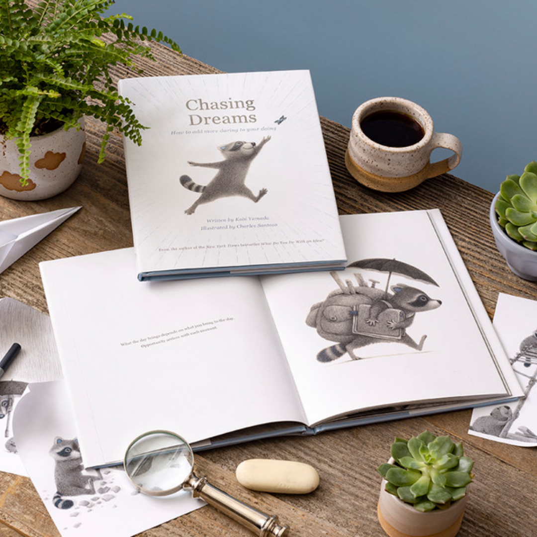 2 copies of "Chasing  Dreams" one open, one closed displayed on a wooden table. The table is decorated with potted plants, a cop of coffee, a magnifying glass, and raccoon drawings.