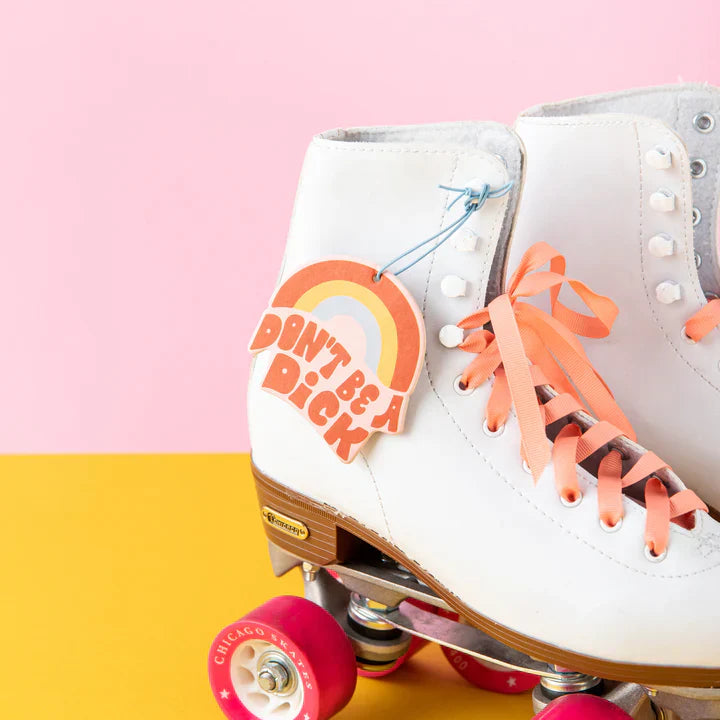 White roller skates with pink wheels and orange laces on a pink and yellow background with don't be a dick air freshener attached.