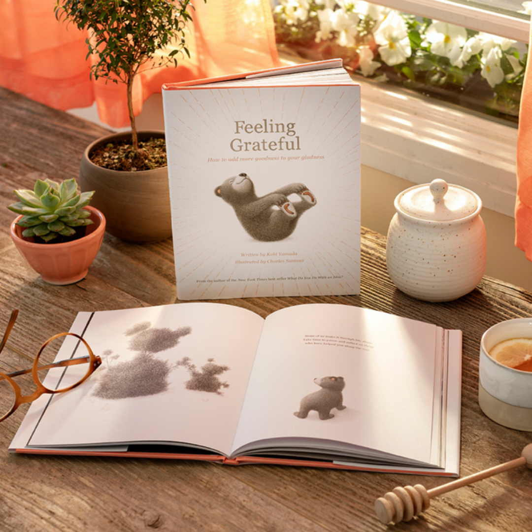 2 copies of "Feeling Grateful" one open, one closed sit on a wood table near a window full of sunlight and flowers. The table is decorated with a pair of plants, a pair of glasses, a honey pot and stirrer, and a cup of tea with lemon.