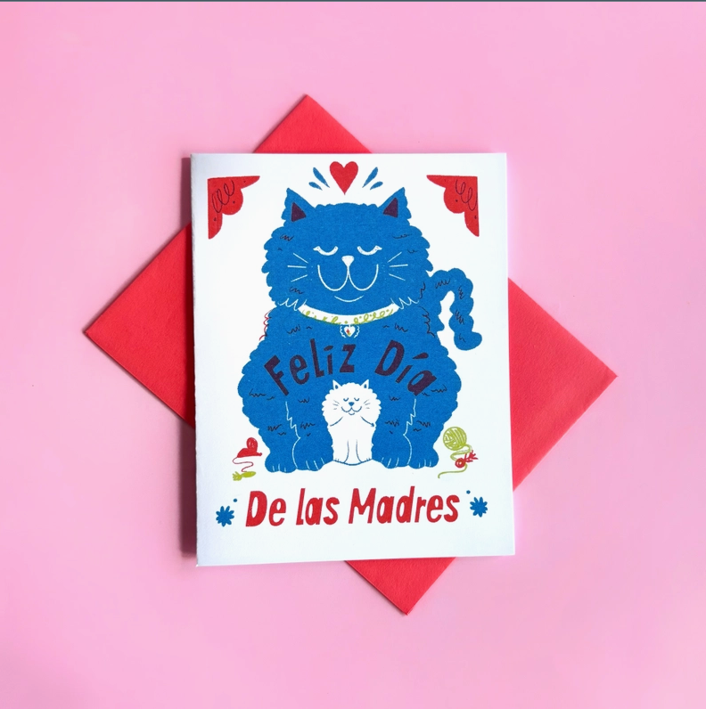White card with red envelope on a pink background. Card features a cartoon blue cat and white kitten with the text "Feliz Dia De Las Madres"
