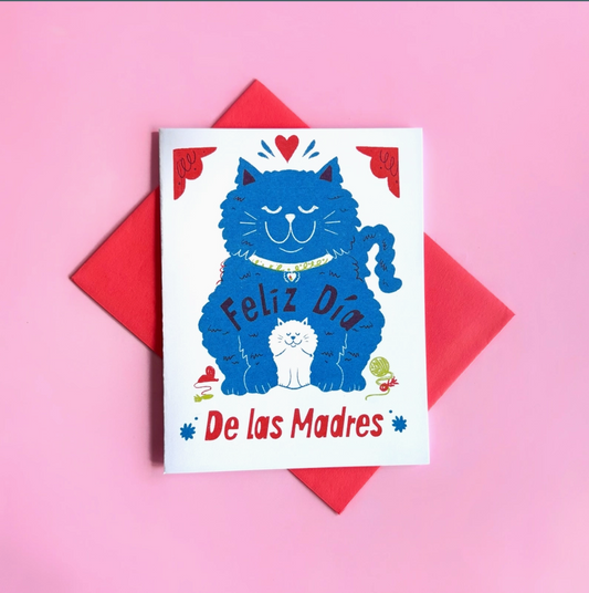 White card with red envelope on a pink background. Card features a cartoon blue cat and white kitten with the text "Feliz Dia De Las Madres"