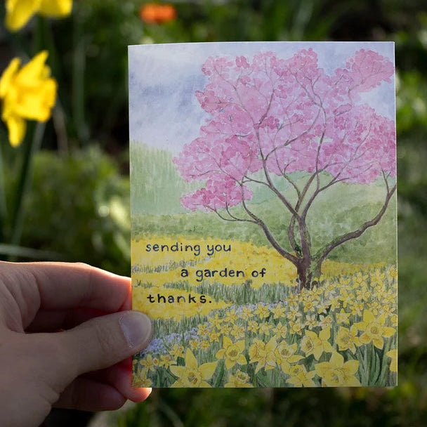 Card with a tree and flowers held in front of a garden with yellow flowers.
