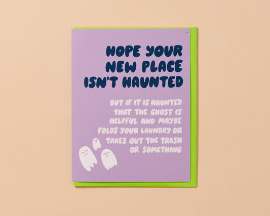 A  greeting card with a lavender front and a bright green envelope, shown standing against a warm beige background. The card reads, 'HOPE YOUR NEW PLACE ISN'T HAUNTED BUT IF IT IS HAUNTED THAT THE GHOST IS HELPFUL AND MAYBE FOLDS YOUR LAUNDRY OR TAKES OUT THE TRASH OR SOMETHING,' with small white ghost illustrations at the bottom.