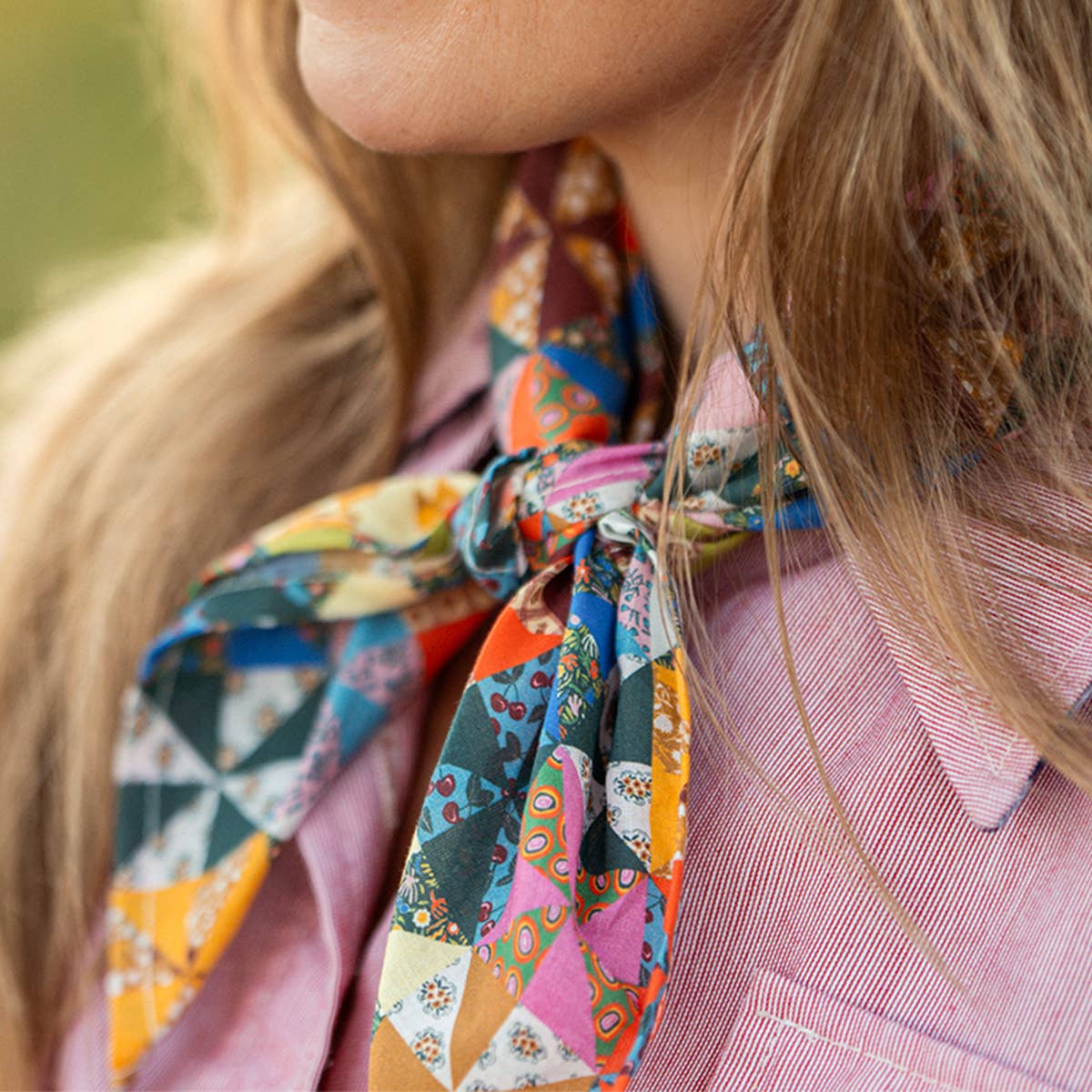 A close-up lifestyle shot of a woman wearing a vibrant, multicolored patchwork-patterned silk scarf tied around her neck, paired with a pink striped button-down shirt.