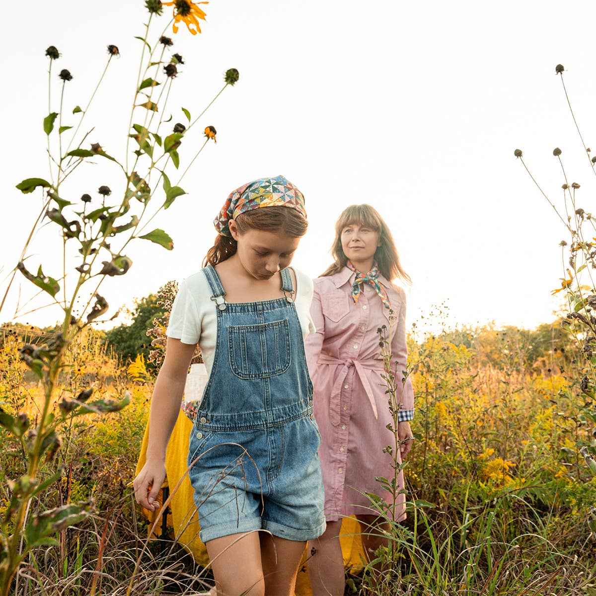 Two women walking outdoors in a sunlit field of wildflowers. One wears denim overall shorts over a white top with a colorful patterned headscarf, and the other wears a dusty pink dress with a matching headscarf.