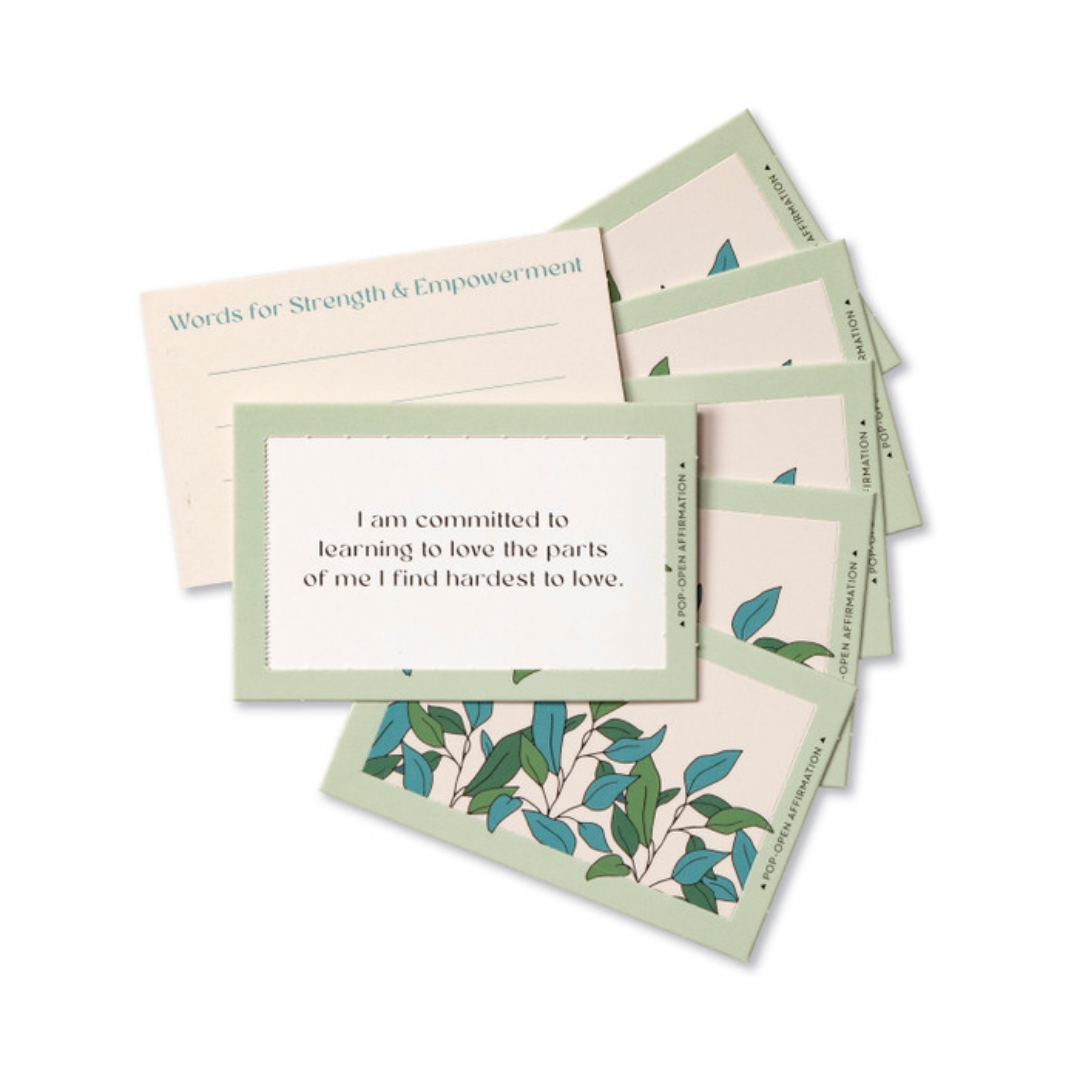 A fan of pop-open affirmation cards. The backs decorated with peaceful looking leaves. The central card reads "I am committed to learning to love the parts of me I find hardest to love," behind it is a card to write your own "Words for Strength & Empowerment"