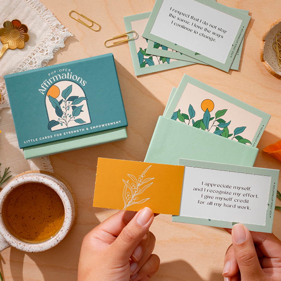 A scene is set at a wood table with the Affirmations and a cup of tea. There's a closed box of cards and an open one, on the table there's a card that reads "I respect that I do not stay the same. I love the ways I continue to change." A pair of hands holds another card that reads "I appreciate myself and I recognize my effort. I give myself credit for all my hard work."