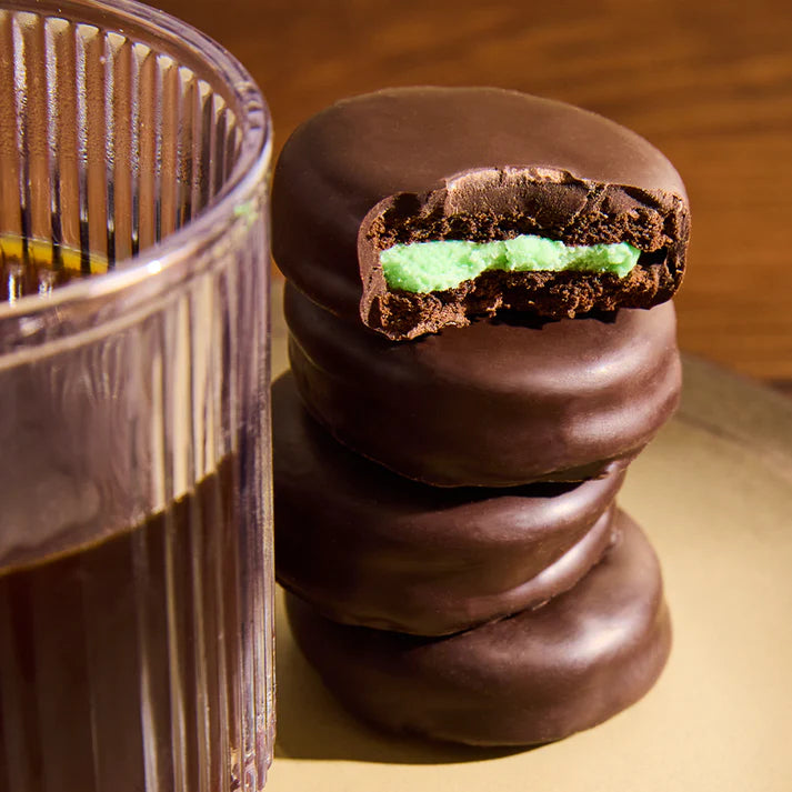 Stack of dark chocolate covered cookies with a mint green filling next to a glass of milk on a wooden surface.