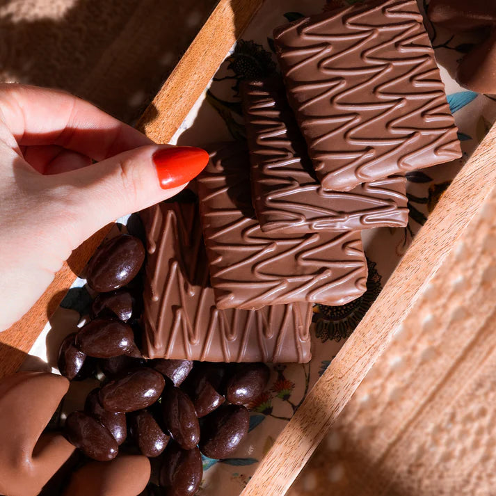 Hand holding a chocolate covered graham cracker, surrounded by more chocolate pieces and almonds on a wooden surface.