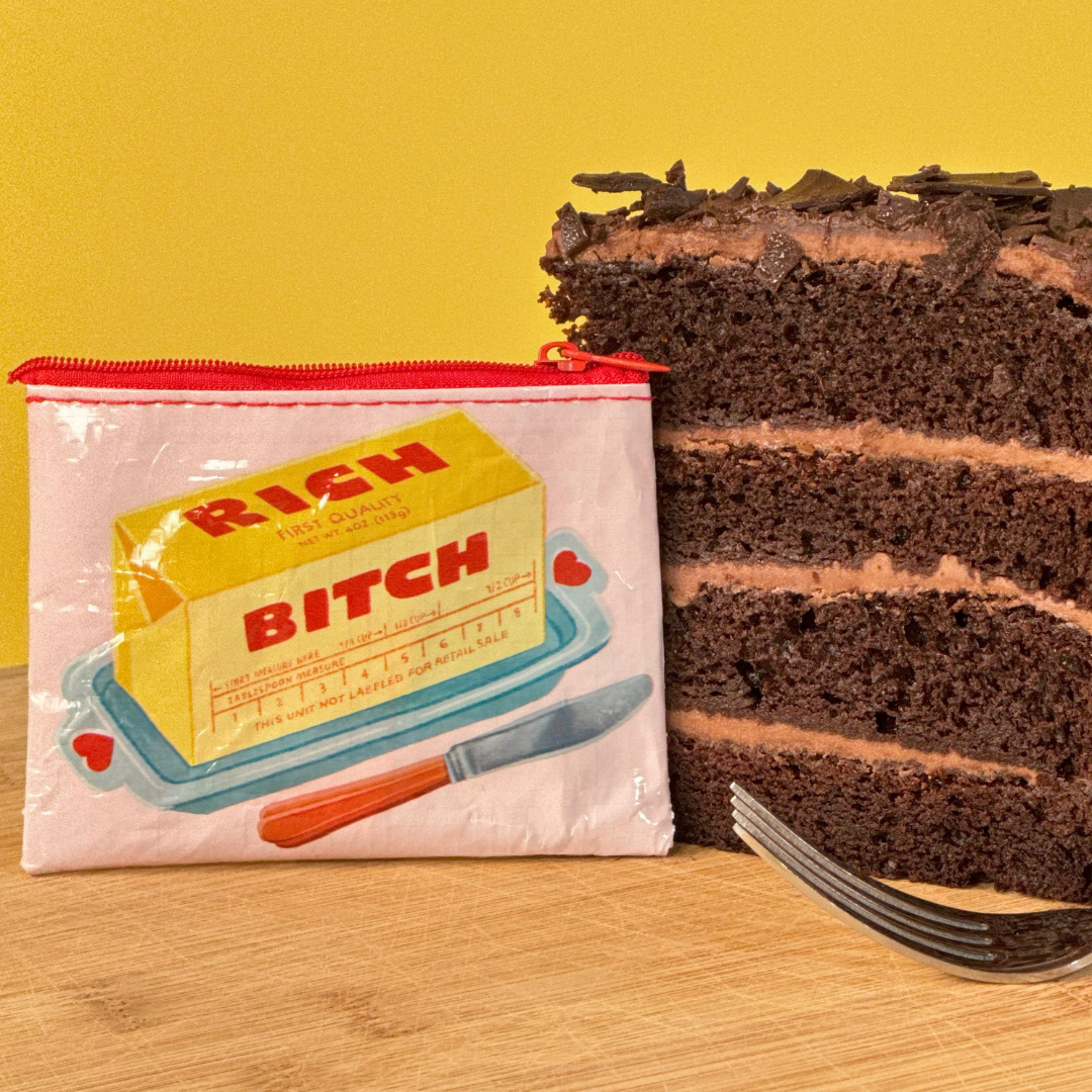 Pink "Rich B*tch" coin purse alongside a very rich looking piece of chocolate 4 layer cake and a fork ready to dig in. 