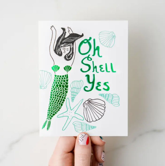 Hand holding white card with illustrated mermaid and shells. Text in green says "Oh Shell Yes"