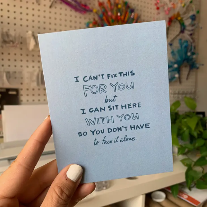Hand holding blue card in front of bright background. Text in blue reads "I can't fix this for you but I can sit here with you so you don't have to face it alone.