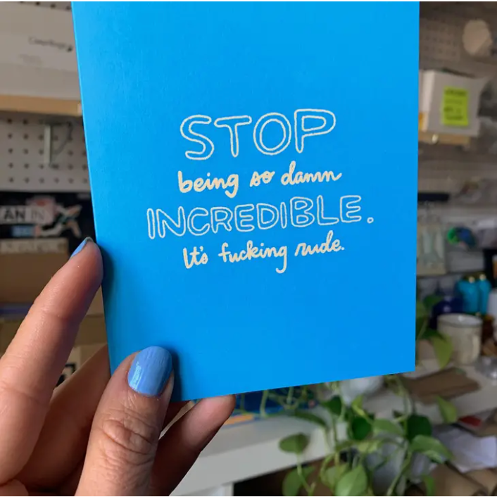 Hand holding blue card in front of blurred background. Text in white reads " Stop being. so damn incredible. It's fucking rude."
