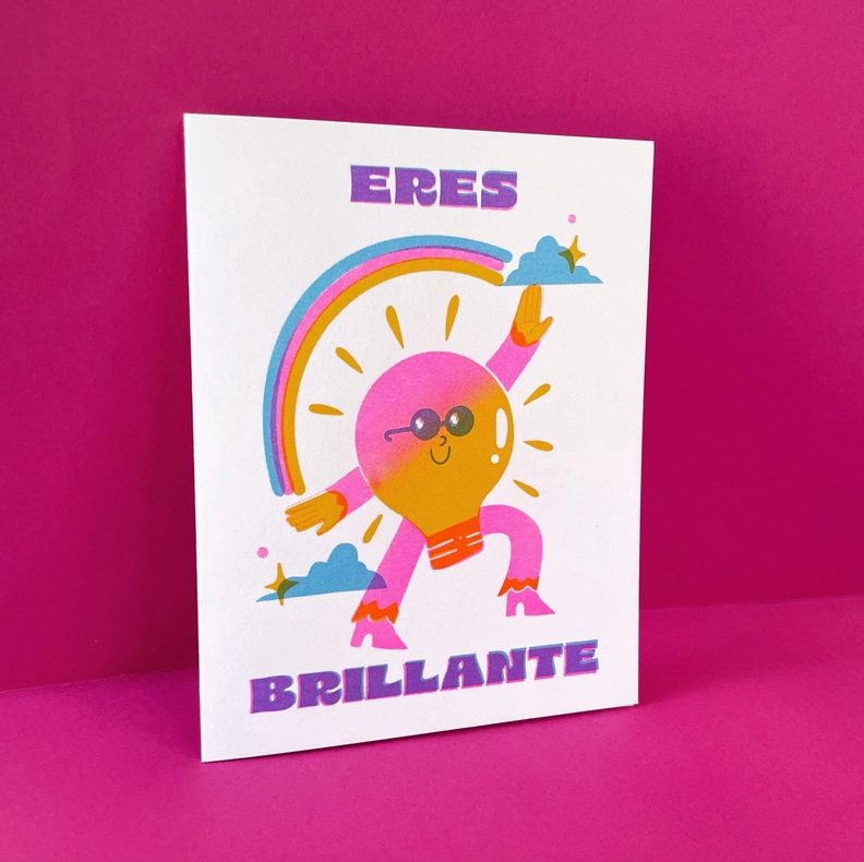 White card on pink background featuring a cartoon rainbow and lightbulb. Text reads in Spanish "Eres Brillante"