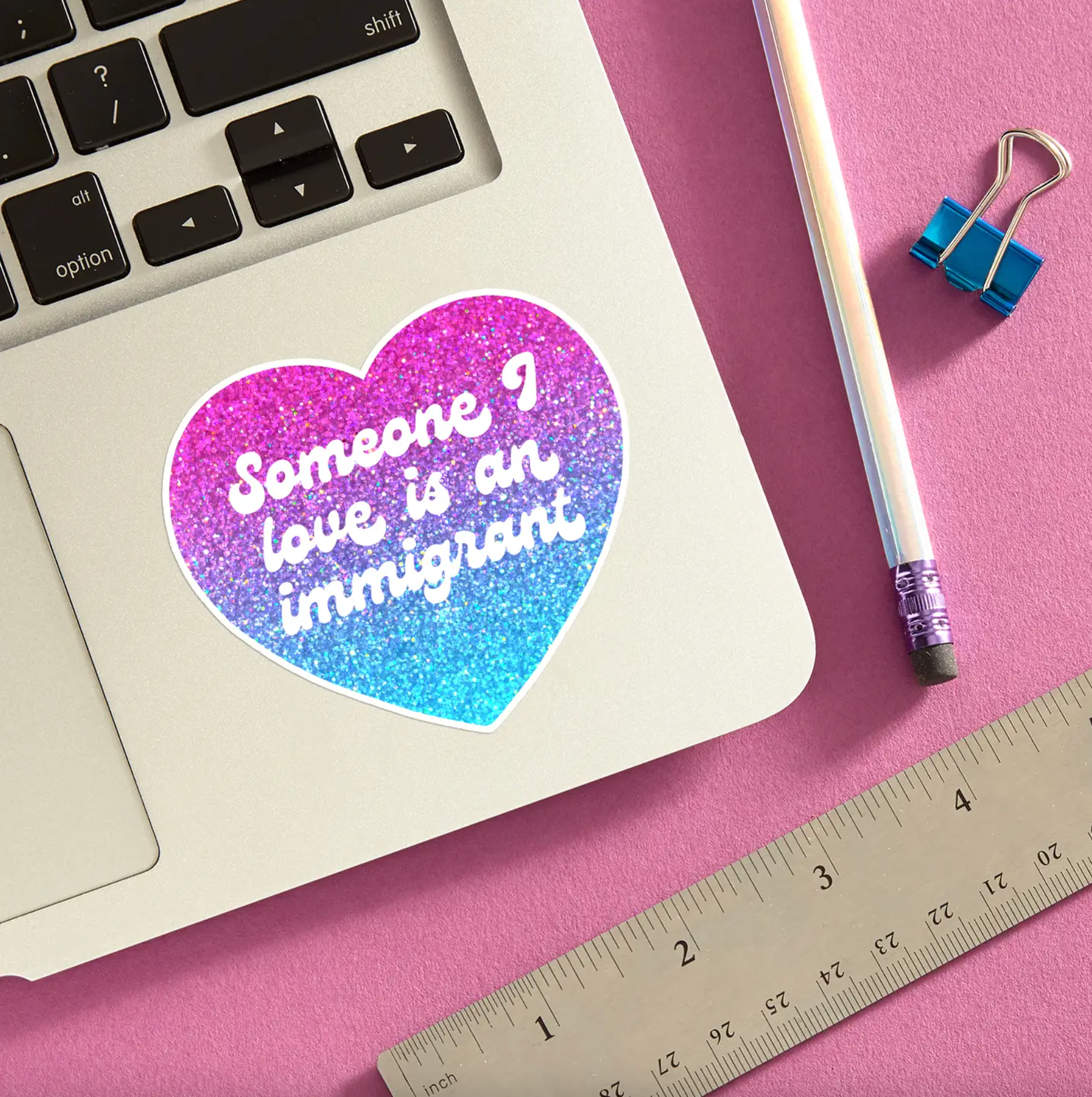 Heart shaped sticker with glitter background gradient form pink to blue. White cursive text that says "Someone I love is an immigrant" place on the keyboard of the laptop.