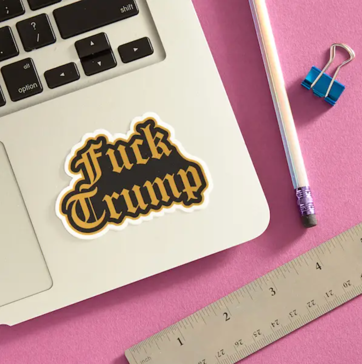 Black and gold "Fuck Trump" sticker on corner of laptop sitting on a pink background.