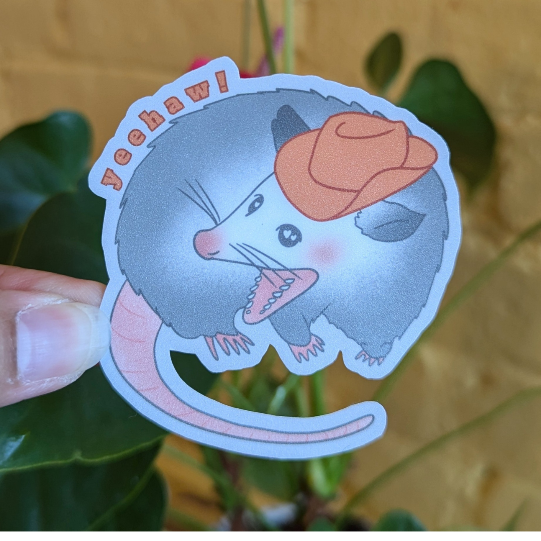 Sticker featuring gray opossum wearing orange cowboy hat and the words "yeehaw!" above it.