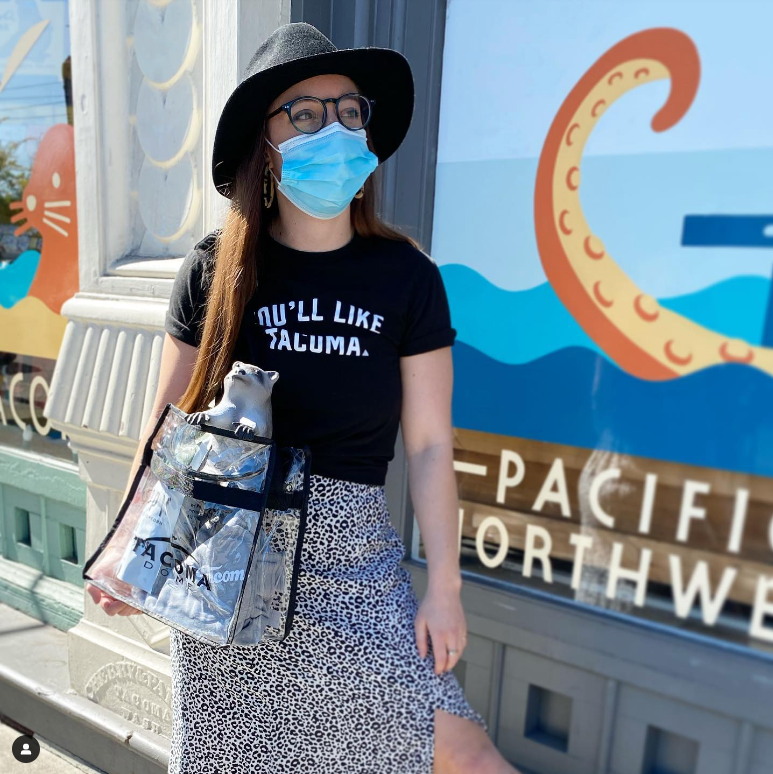 Person wearing a black t-shirt with text, holding a clear bag with a small animal design, standing in front of a colorful mural.
