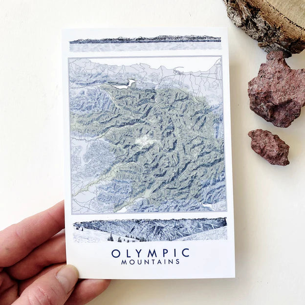 Map of Olympic Mountains on a postcard that says OLYMPIC MOUNTAINS, held by a hand with rocks on a white background