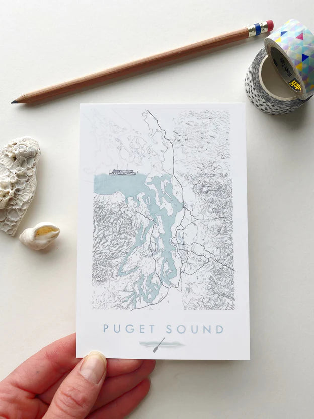 Postcard with a map of Puget Sound held by a hand on a white surface with a pencil and tape measure.