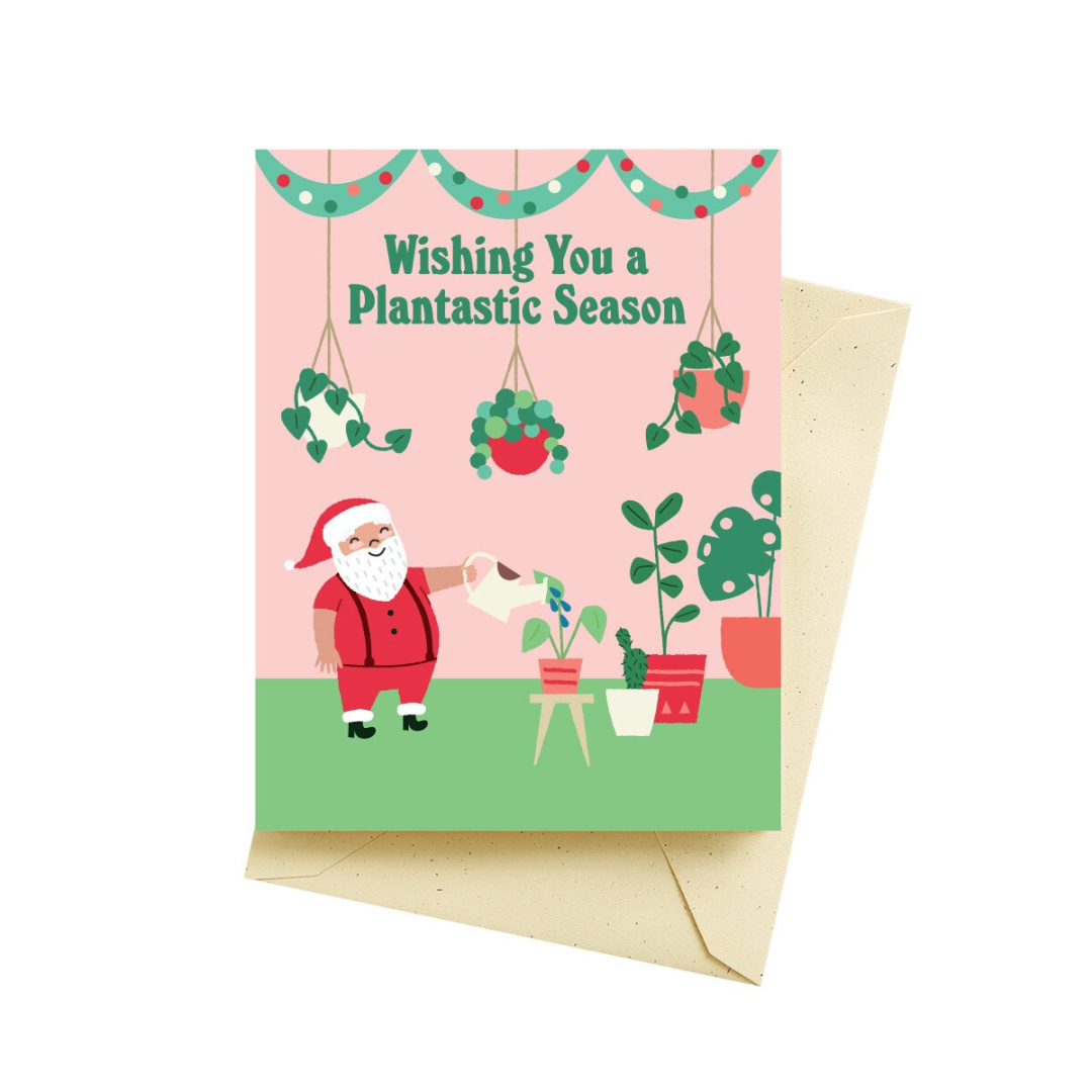 Holiday greeting card with pink background and Santa watering plants featuring the words "Wishing you a plantastic season" in a green font. Pictured with a tan colored envelope.