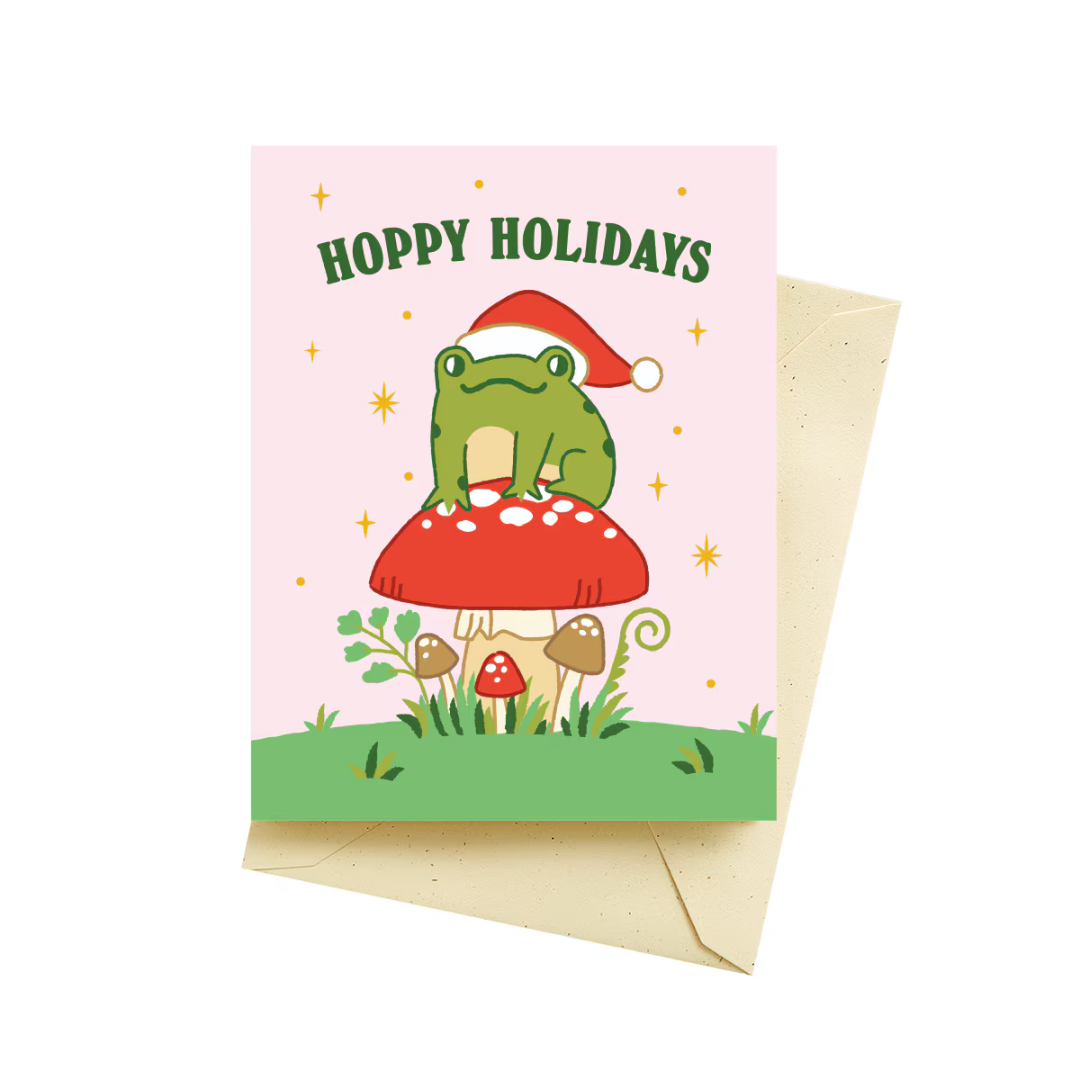 Baby pink holiday card featuring a green Christmas frog wearing a red Santa hat, sitting on top of a red and white mushroom surrounded by more red and brown mushrooms and green grass. Gold stars twinkle around the frog along with the words "hoppy holidays" in a darker green. Featured with a tan colored envelope.