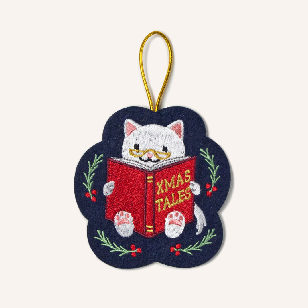 Felt navy blue Christmas ornament featuring a white cat reading a book titled "xmas tales" surrounded by holly, strung with gold cord.