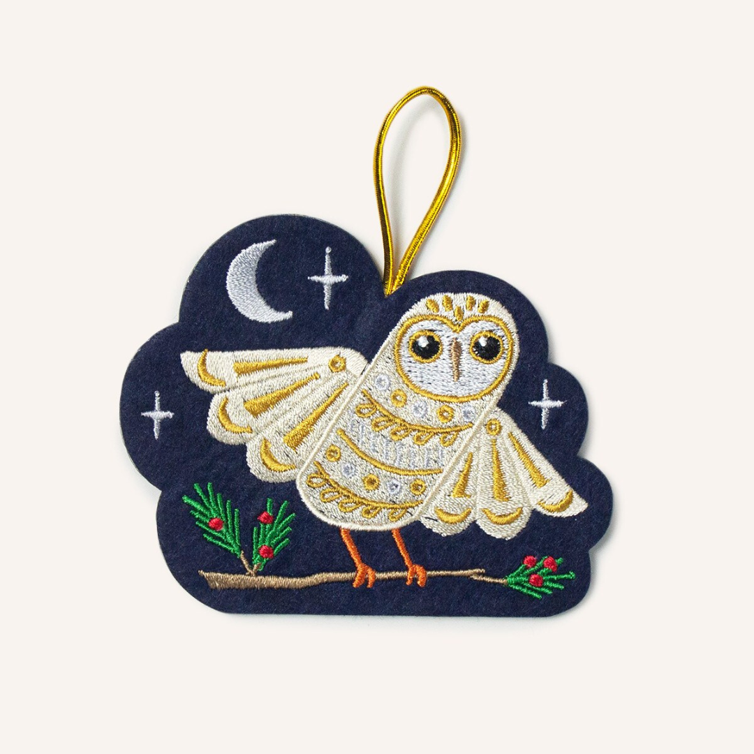 Navy blue felt ornament featuring a white and gold owl perched on a branch of holly surrounded by silver stars and a moon. Hung by gold cord.