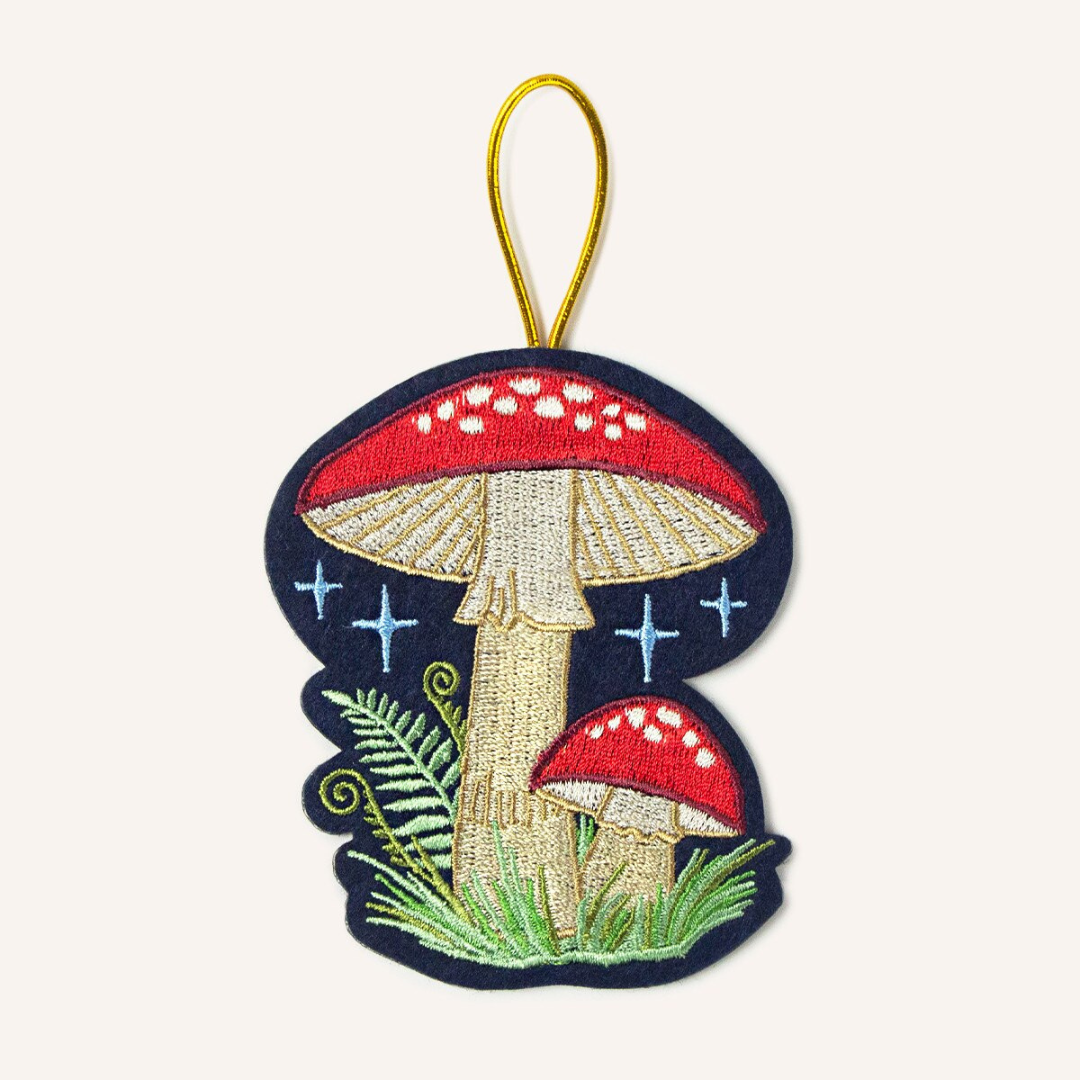 Navy blue felt ornament featuring a red and white mushroom with green ferns at the base of it, strung by gold cord.