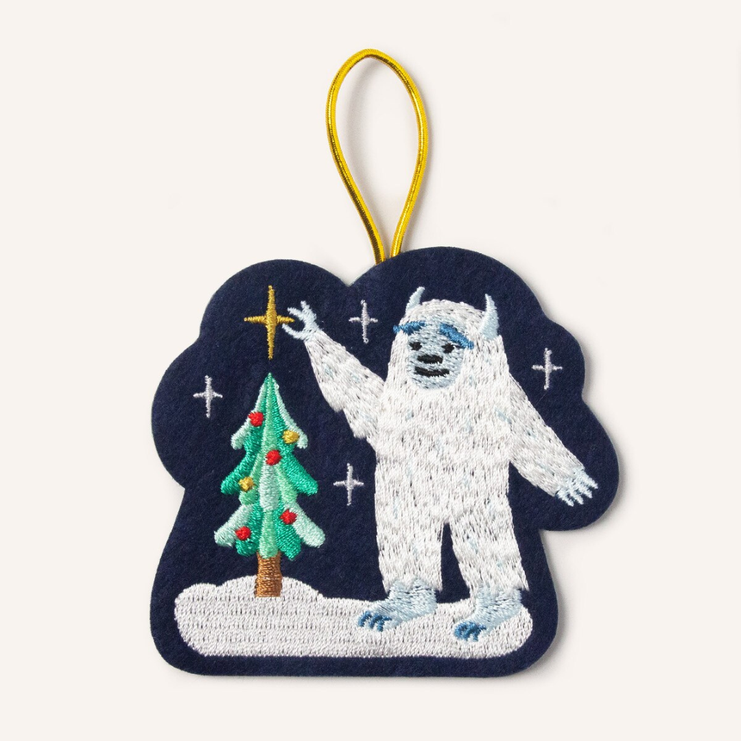 Navy blue felt ornament featuring a white yeti standing in the snow, putting a gold star on top of a Christmas tree. Hung by a gold cord.