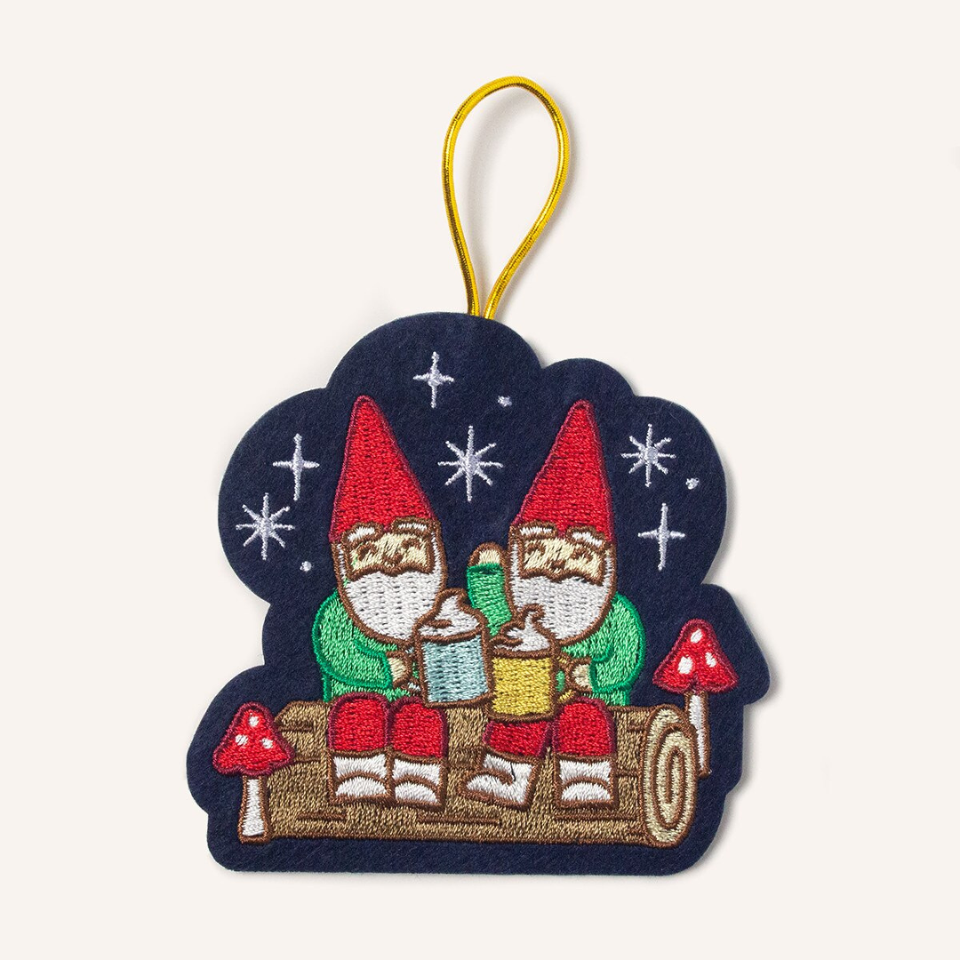 Navy blue felt ornament strung with gold cord featuring two gnomes sitting on a brown log drinking hot chocolates together surrounded by red mushrooms and silver stars.