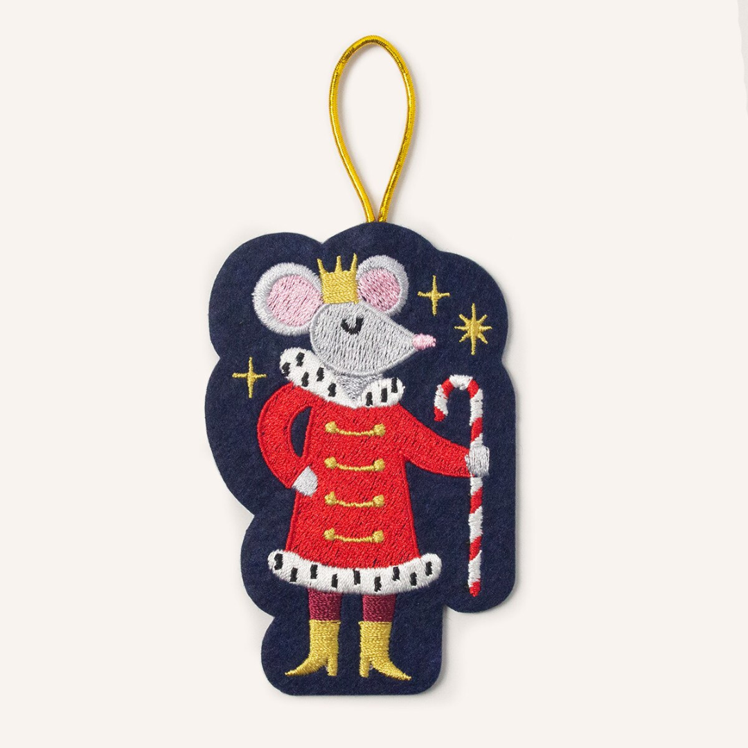 Navy blue felt ornament strung by gold cord featuring a rat wearing a gold crown and boots with a red coat holding a red and white striped candy cane, surrounded by gold stars.