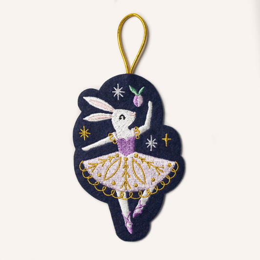 Navy blue felt ornament hung by a gold cord featuring a white rabbit ballerina in a purple and gold tutu dress surrounded by sugarplums and stars.