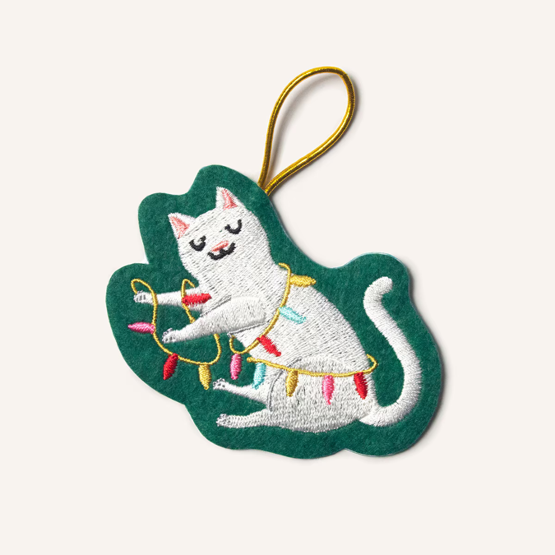 A wintergreen felt ornament featuring a white cat tangled in pink, red, yellow and blue Christmas lights. Hung by a gold cord.