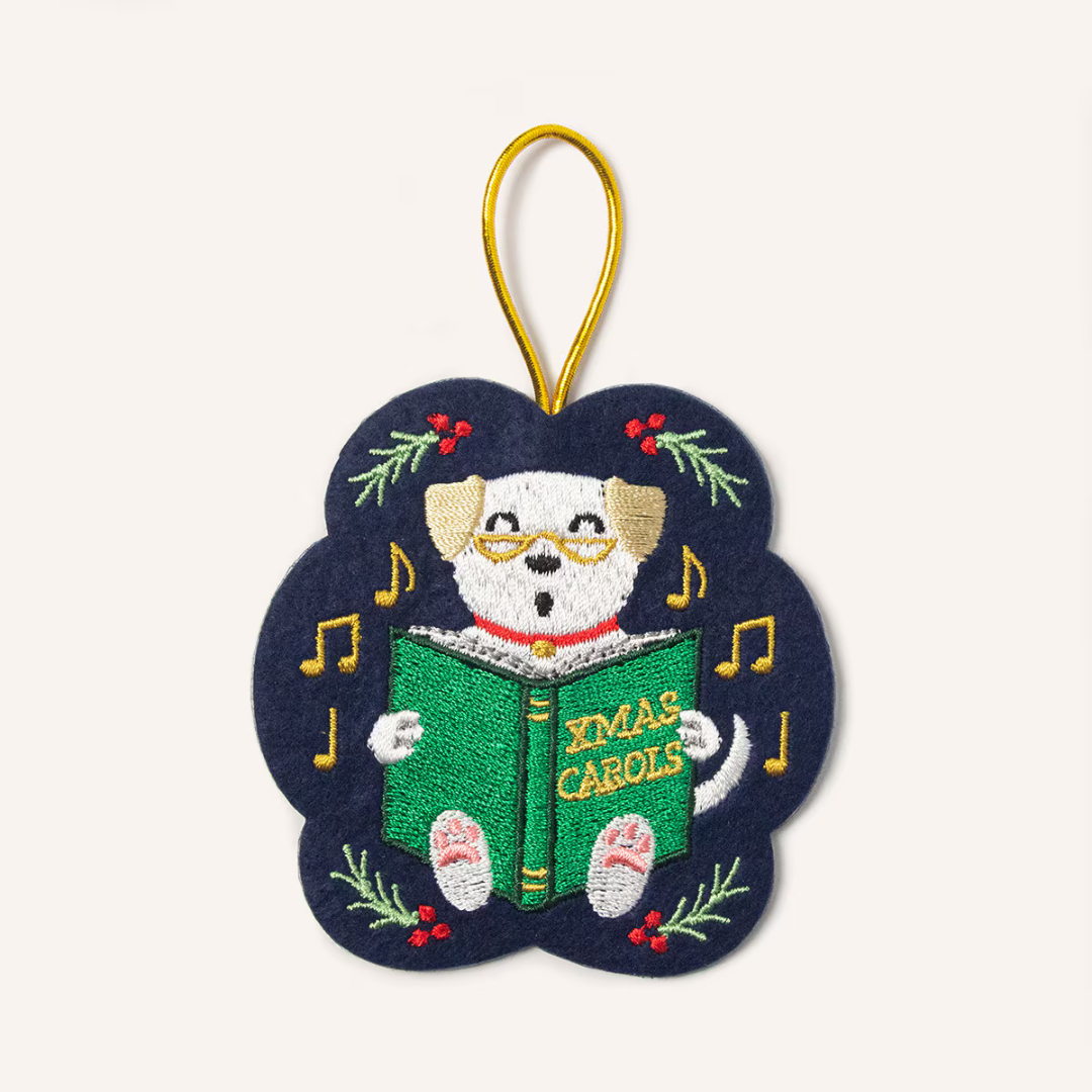 Navy blue felt ornament hung by a gold cord featuring a white singing dog wearing glasses, holding a green book titled "Xmas Carols" surrounded by holly and gold musical notes.