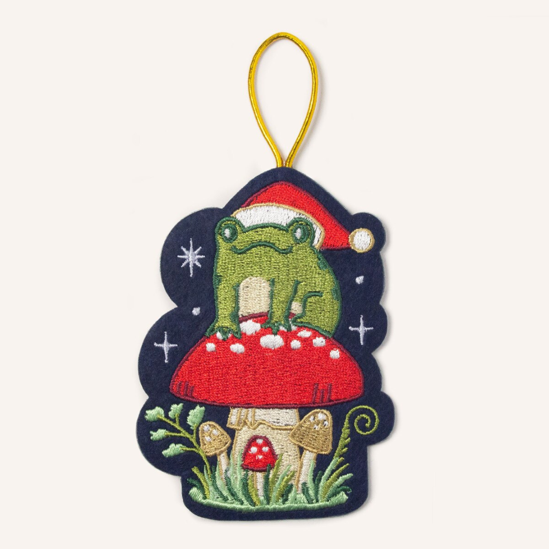 Navy blue felt ornament hung by gold cord featuring a green frog wearing a red santa hat, sitting on top of a red mushroom surrounded by silver stars.