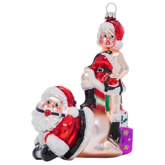 Christmas ornament features a naughty Mrs. Claus standing behind Santa with one leg atop of his back. 