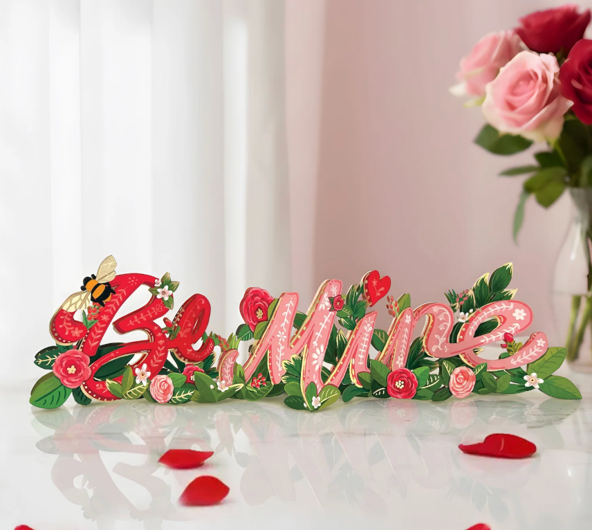 Decorative card with "Be Mine" in pink and red letters, surrounded by flowers and leaves on a white surface.