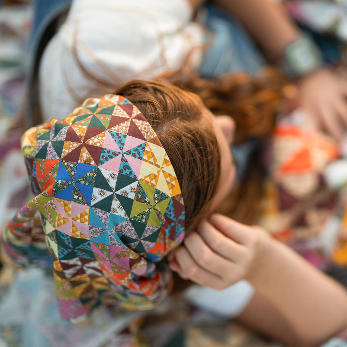 A close-up lifestyle shot of a person wearing a colorful geometric patterned headscarf. The headscarf features a patchwork-style design with triangles in shades of blue, green, orange, pink, yellow, and brown.