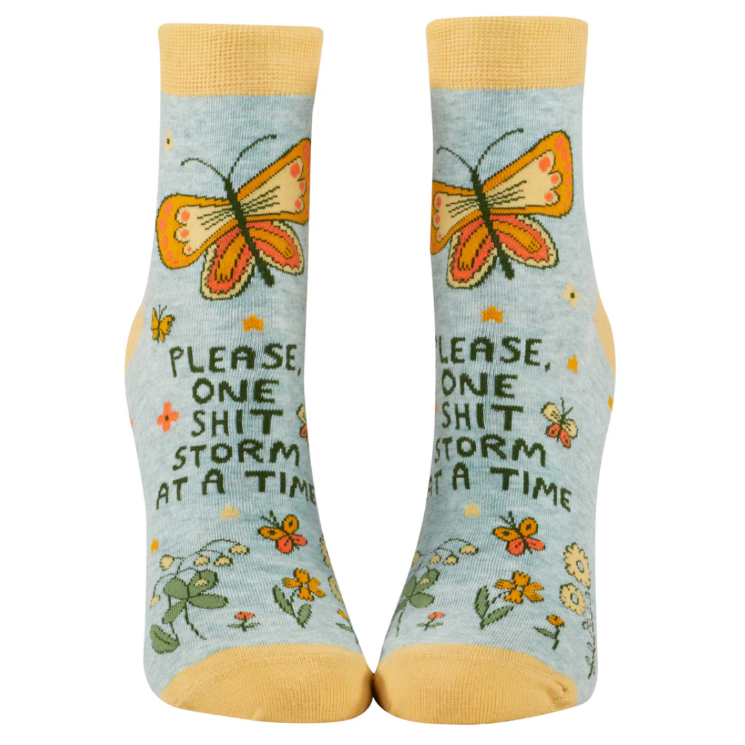 A pair of blue ankle socks with a butterfly and floral design, featuring the phrase 'PLEASE, ONE SHITSTORM AT A TIME'.