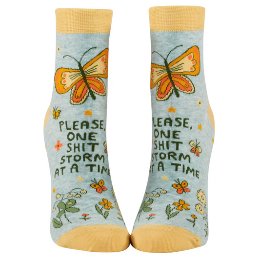 A pair of blue ankle socks with a butterfly and floral design, featuring the phrase 'PLEASE, ONE SHITSTORM AT A TIME'.