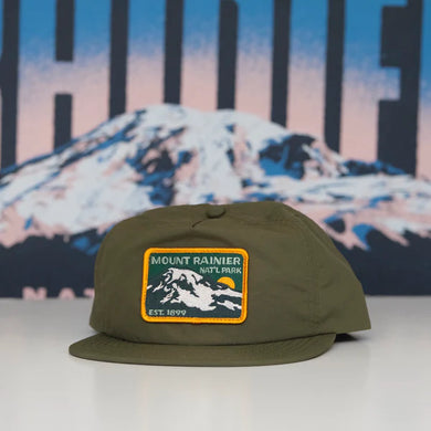 green hat with mount rainier patch on front sits ontable with picture of mount rainier in background